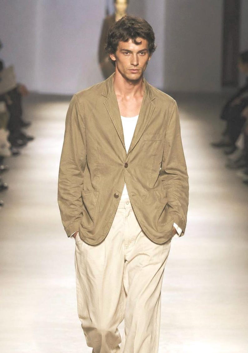 May include: A tan blazer with a single button closure, paired with cream-colored pants and a white undershirt. The outfit is worn by a person on a runway. The blazer has a classic, tailored fit.