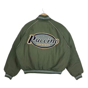 May include: An olive green bomber jacket featuring a large oval patch on the back with the text Paccino and the phrase THIS IS JUST RIGHT FOR YOU. The jacket has a ribbed collar and cuffs with grey stripes and white piping along the shoulders.