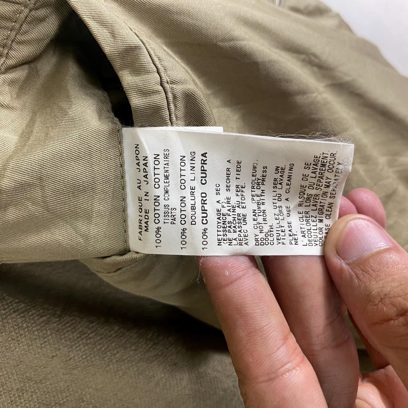 May include: Close-up of a beige fabric tag with text in English and French. The tag reads "FABRIQUE AU JAPON MADE IN JAPAN" and details the fabric composition as 100% cotton and 100% cupro lining. Care instructions are also printed on the tag.