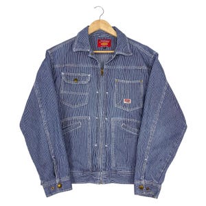 May include: A blue and white hickory stripe zip-up jacket featuring a fold-down collar, long sleeves with buttoned cuffs, and four front pockets. The jacket includes a red brand label at the inner neck and a small rectangular logo on the chest pocket.