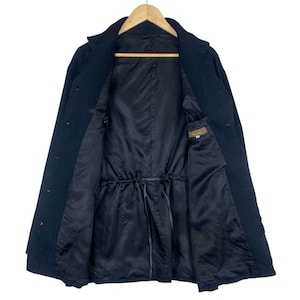 K&ouml;nnte beinhalten: Ein schwarzer Mantel mit Knopfleiste und Kordelzug in der Taille. Der Mantel ist offen und zeigt ein schwarzes Futter und eine kleine Tasche. Der Mantel h&auml;ngt an einem Holzb&uuml;gel.