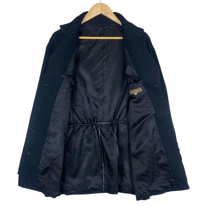 K&ouml;nnte beinhalten: Ein schwarzer Mantel mit Knopfleiste und Kordelzug in der Taille. Der Mantel ist offen und zeigt ein schwarzes Futter und eine kleine Tasche. Der Mantel h&auml;ngt an einem Holzb&uuml;gel.