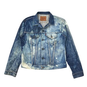 May include: A faded blue denim jacket with a distressed appearance and white paint marks. It has a button-up front, chest pockets, and a small red tag. The brand name "EDWIN" is displayed on a brown leather patch.