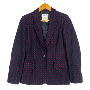 Op de afbeelding: Marineblauwe blazer met een rood stippenpatroon. De blazer heeft een enkele knoop, twee voorzakken en een label met de tekst "Bellezza". Hij hangt aan een houten hanger.