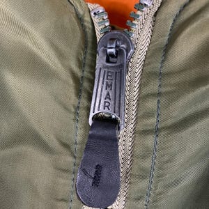 Puede incluir: Primer plano de una cremallera de chaqueta verde oliva con la palabra "EMAR" impresa en el tirador met&aacute;lico. La cremallera est&aacute; parcialmente cerrada, revelando un forro naranja. El tirador es de cuero negro.