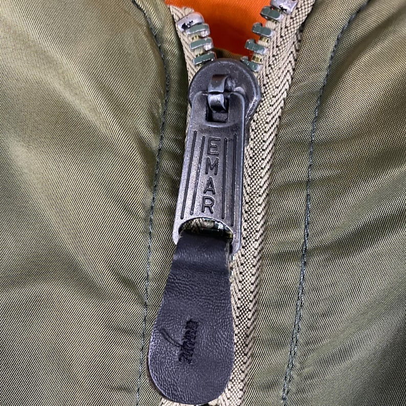 Puede incluir: Primer plano de una cremallera de chaqueta verde oliva con la palabra "EMAR" impresa en el tirador met&aacute;lico. La cremallera est&aacute; parcialmente cerrada, revelando un forro naranja. El tirador es de cuero negro.