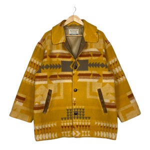 May include: A mustard yellow jacket featuring a geometric pattern in shades of brown, cream, and gray. The jacket has a wide collar, three dark buttons down the front, and two vertical pockets with brown trim. A label is visible inside the collar.