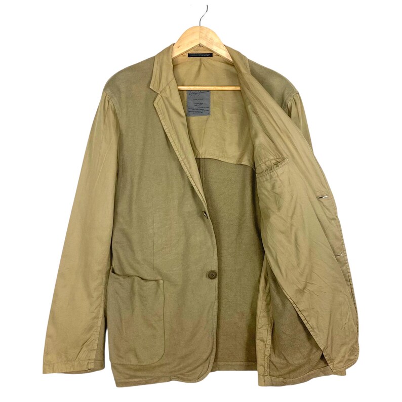 May include: A tan blazer with a single-button closure, two front pockets, and a notched lapel. The jacket is hanging on a wooden hanger against a white background. The interior lining is a lighter shade of tan.