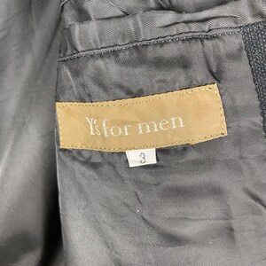 K&ouml;nnte beinhalten: Nahaufnahme des grauen Innenfutters einer Jacke mit einem beigefarbenen Etikett mit der Aufschrift "Y's for men". Unter dem Etikett befindet sich die wei&szlig;e Zahl "3". Der Stoff der Jacke ist strukturiert.
