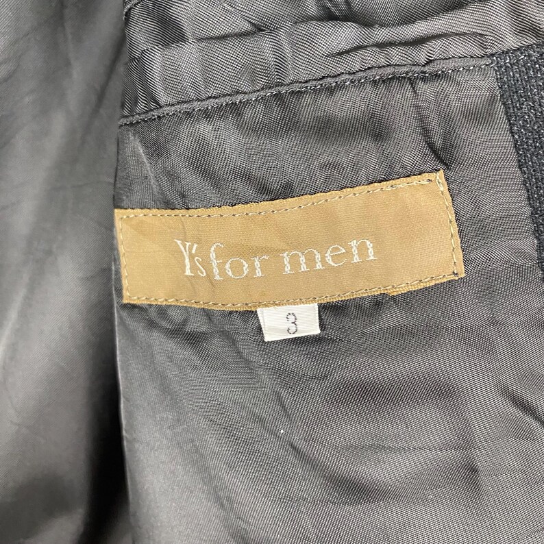 K&ouml;nnte beinhalten: Nahaufnahme des grauen Innenfutters einer Jacke mit einem beigefarbenen Etikett mit der Aufschrift "Y's for men". Unter dem Etikett befindet sich die wei&szlig;e Zahl "3". Der Stoff der Jacke ist strukturiert.