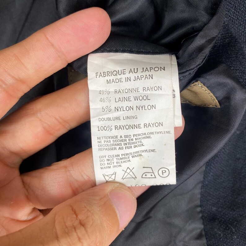 K&ouml;nnte beinhalten: Nahaufnahme eines Kleidungsst&uuml;cketiketts mit Text in Franz&ouml;sisch und Englisch. Das Etikett lautet "FABRIQUE AU JAPON MADE IN JAPAN" und gibt die Stoffzusammensetzung an, einschlie&szlig;lich Viskose, Wolle und Nylon. Pflegehinweise sind ebenfalls enthalten.