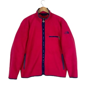Può includere: Una giacca in pile rosa acceso con cerniera frontale integrale blu scuro, collo alto e due tasche laterali. Sul lato sinistro è presente un piccolo taschino sul petto con cerniera e il logo The North Face ricamato in blu scuro.