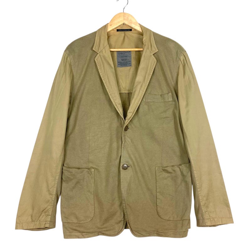 May include: A tan blazer with a two-button closure, two front pockets, and a notched lapel. The jacket is hanging on a wooden hanger. The sleeves are a lighter shade of tan than the body of the jacket.