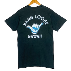 May include: Black t-shirt with a white and blue hand gesture graphic. The text "HANG LOOSE" arches above the hand, and "HAWAII" is printed below. The shirt is hanging on a wooden hanger.