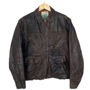 May include: A brown leather jacket with a collared neckline, a zipper closure, and multiple pockets. The jacket is hanging on a wooden hanger. The leather has a worn, textured appearance.