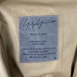 May include: Close-up of a tan fabric with a rectangular label. The label is light blue with the handwritten signature "Yohji Yamamoto" and the text "POUR HOMME". Additional text includes "FABRIQUE AU JAPON", "MADE IN JAPAN", and washing instructions.