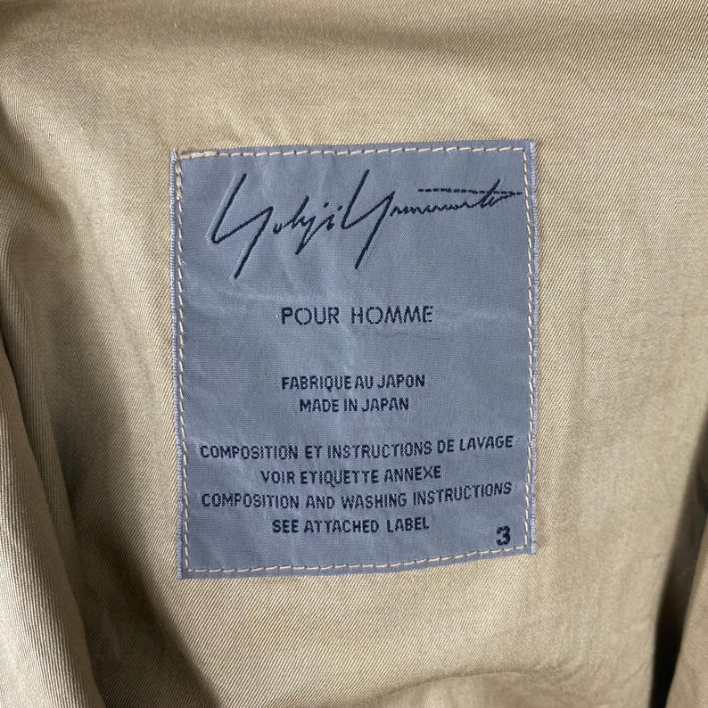 May include: Close-up of a tan fabric with a rectangular label. The label is light blue with the handwritten signature "Yohji Yamamoto" and the text "POUR HOMME". Additional text includes "FABRIQUE AU JAPON", "MADE IN JAPAN", and washing instructions.