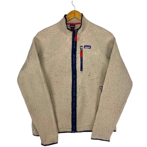 Può includere: Giacca in pile beige con cerniera a tutta lunghezza e collo alto. La giacca presenta finiture blu navy lungo la cerniera e il colletto, con un tirazip rosso. Una piccola toppa con un logo è sul petto.