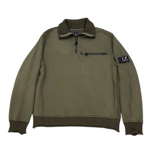 May include: An olive green long-sleeve pullover sweatshirt featuring a ribbed collar, cuffs, and hem. It includes a quarter-zip neckline, a horizontal zippered chest pocket, and a black C.P. Company brand patch on the upper left sleeve.