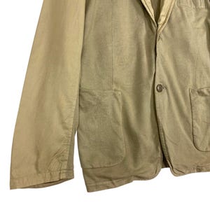 May include: A tan blazer with a single-button closure and two front pockets. The jacket has a notched lapel and long sleeves. The fabric appears to be a lightweight cotton or linen blend, suitable for warmer weather.