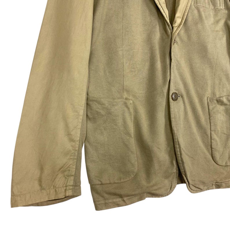 May include: A tan blazer with a single-button closure and two front pockets. The jacket has a notched lapel and long sleeves. The fabric appears to be a lightweight cotton or linen blend, suitable for warmer weather.