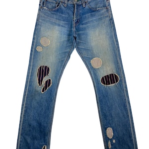 May include: Light blue denim jeans with distressed details, including patches and rips. The jeans are hanging on a wooden hanger. The jeans have a classic five-pocket design and a button and zip closure.