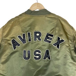 Puede incluir: Chaqueta bomber verde oliva con el texto "AVIREX USA" en letras negras y blancas en la espalda. La chaqueta tiene cuello y pu&ntilde;os de canal&eacute;. La chaqueta est&aacute; colgada de una percha de madera.