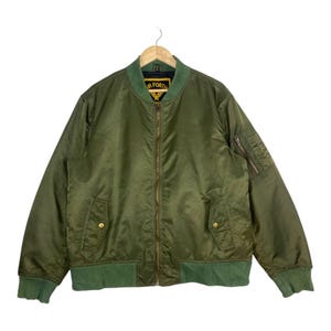 Peut inclure: Veste de bombardier vert olive avec fermeture éclair, deux poches avant et une petite poche sur la manche gauche. La veste a un col, des poignets et un ourlet côtelés verts. La veste est suspendue à un cintre en bois.