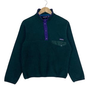 Puede incluir: Un suéter de forro polar verde oscuro con cuello alto y una tapeta con botones a presión de color morado. La prenda incluye un bolsillo en el pecho con solapa y una pequeña etiqueta de la marca Patagonia en el lado derecho, sobre una percha.