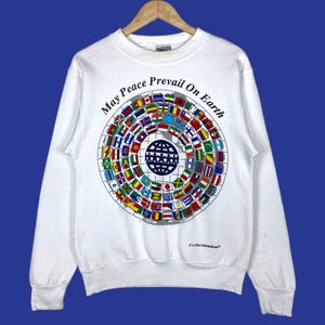 May include: White sweatshirt with a crew neck and long sleeves. The front features a circular design with flags of various countries surrounding a globe graphic. The text "May Peace Prevail On Earth" is arched above the design.