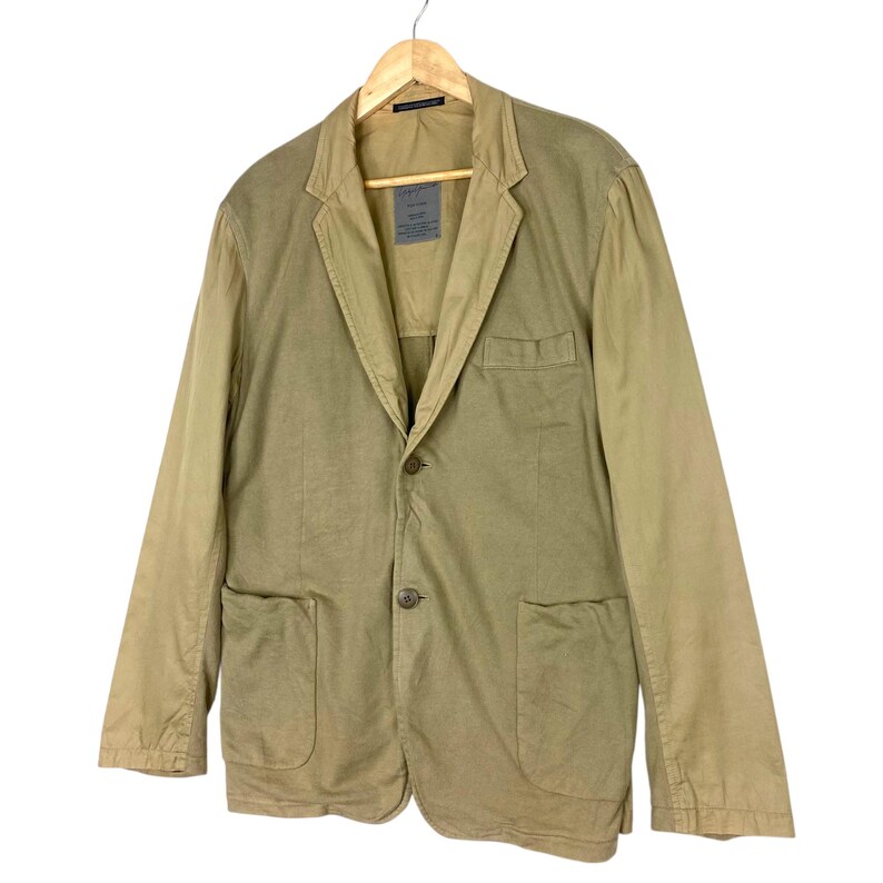 May include: A tan blazer with two front pockets, a single button closure, and a notched lapel. The jacket is hanging on a wooden hanger against a white background. The sleeves are a lighter shade of tan than the body of the jacket.