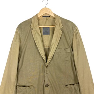 May include: A two-tone blazer in shades of tan and beige, hanging on a wooden hanger. The jacket features a notched lapel, two front pockets, and a single-button closure. A label is visible on the chest. The garment is made of a lightweight fabric.