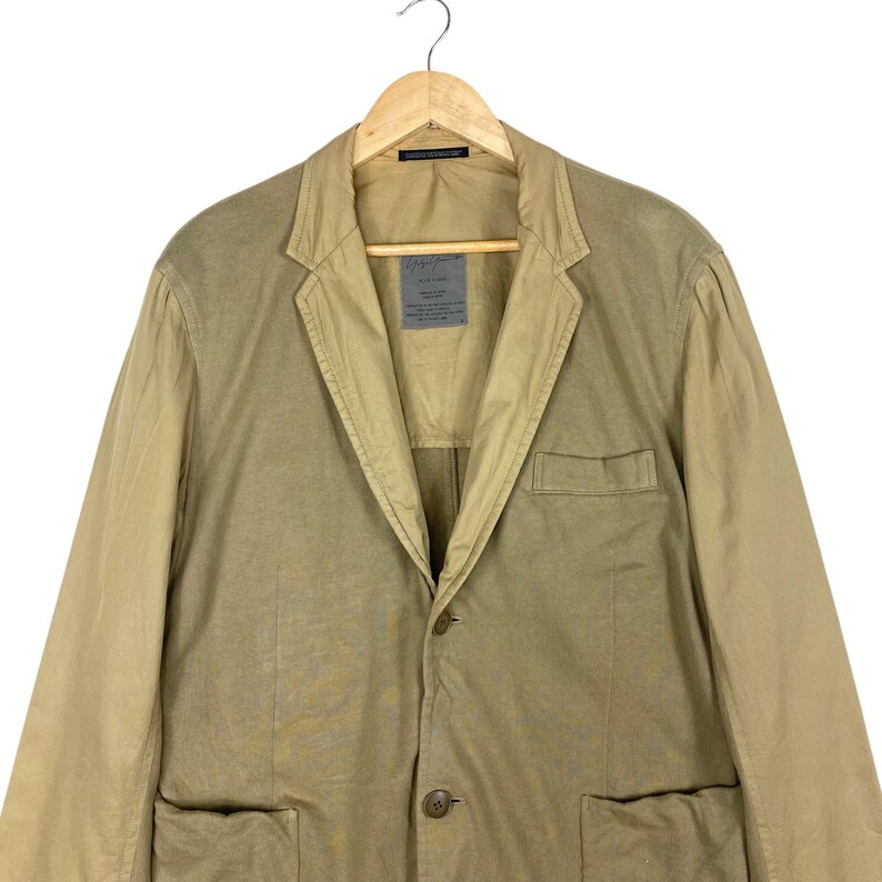 May include: A two-tone blazer in shades of tan and beige, hanging on a wooden hanger. The jacket features a notched lapel, two front pockets, and a single-button closure. A label is visible on the chest. The garment is made of a lightweight fabric.
