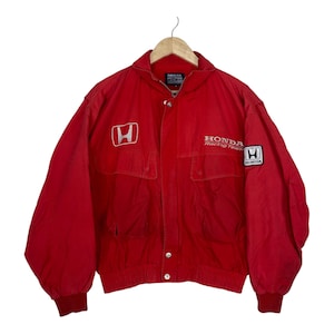 May include: A bright red Honda Racing Team jacket featuring a high collar, long sleeves with ribbed cuffs, and a front button closure. The chest displays the white Honda logo and text, with an additional Honda patch on the left sleeve.
