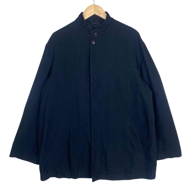 K&ouml;nnte beinhalten: Schwarze Jacke mit Stehkragen und zwei Vordertaschen. Die Jacke hat einen Knopfverschluss und lange &Auml;rmel. Das Kleidungsst&uuml;ck h&auml;ngt an einem Holzb&uuml;gel vor wei&szlig;em Hintergrund.