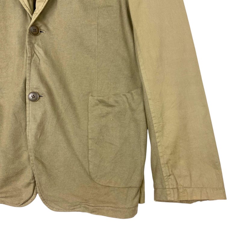 May include: Close-up of a tan blazer with two brown buttons and a front pocket. The blazer has a classic design with a notched lapel and long sleeves. The fabric appears to be a lightweight cotton or linen blend, suitable for warmer weather.