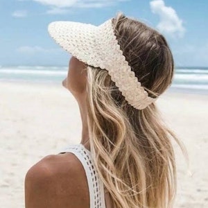 May include: A woven straw visor in a natural beige color. The visor has a scalloped edge and is worn on the head. The image shows the visor from the side, with the beach and ocean in the background. The visor is a summer accessory.
