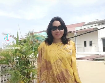 Puede incluir: Una mujer con gafas de sol y un vestido de manga larga con estampado amarillo y marrón. El vestido tiene un corte holgado y un diseño fluido.