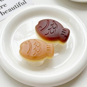 May include: Two fish-shaped desserts, one light brown and one dark brown, sit on a white plate. The desserts have intricate designs and are placed close together. The plate is white with a simple, elegant design.