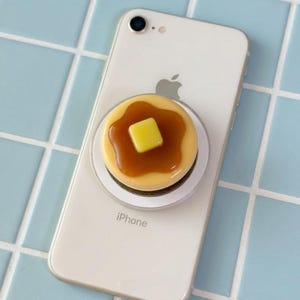 May include: A white iPhone with a pancake-shaped phone grip attached. The grip features a yellow square of butter on top of a pancake covered in syrup. The phone is on a light blue tiled surface.