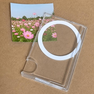 May include: A clear, rectangular phone case featuring a prominent white circular magnetic ring on the back. A square photograph of a vibrant field of pink cosmos flowers is positioned partially underneath the transparent protective cover.