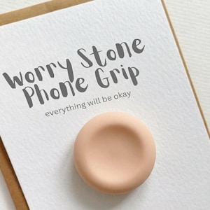 Könnte beinhalten: Ein pfirsichfarbener Handyhalter mit konkavem Zentrum liegt auf einer weißen Karte. Auf der Karte stehen die Worte "Worry Stone Phone Grip" und "everything will be okay" in Grau. Die Karte befindet sich auf einem braunen Hintergrund.