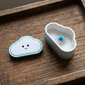 May include: A light blue cloud-shaped case with a smiling face next to a matching container holding a blue mechanical keyboard switch. The items are on a wooden tray.