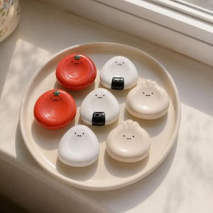 May include: A collection of seven ceramic chopstick rests arranged on a round cream-coloured plate. The set includes two red tomato-shaped rests, two white onigiri-shaped rests with black seaweed details, and three beige dumpling-shaped rests.