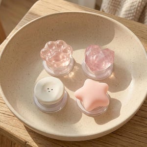 Puede incluir: Un cuenco de cerámica beige contiene cuatro agarres decorativos para teléfono. Los agarres tienen forma de huella de pata, corazón, estrella y cara sonriente. La huella y el corazón son de color rosa translúcido, la estrella es rosa y la cara es blanca.