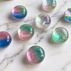 May include: A selection of iridescent, spherical glass beads. Each bead displays a gradient of colours, including blue, pink, green, and purple, with a shimmering, glittery effect. The beads are set against a white marble background.
