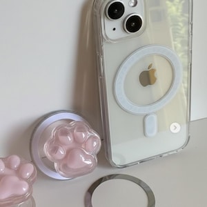 Könnte beinhalten: Transparente Handyhülle mit weißem MagSafe-Ring und einem rosa Pfotenabdruck-Handygriff. Die Handyhülle hat ein klares Design, und der Pfotenabdruck-Griff ist hellrosa. Ein silberner Ring ist ebenfalls enthalten.