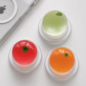 May include: Three spherical, translucent paperweights or decorative objects resting on clear circular bases. One is bright red with a green stem, one is pale green with a single leaf, and one is vibrant orange with a small green stem detail.