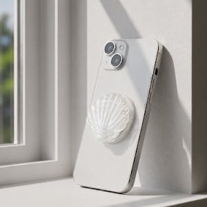 May include: A white smartphone with a dual-camera system on the back, featuring a translucent, iridescent seashell-shaped phone grip attached to the center. The device is positioned upright, displaying a glossy finish and a textured, ridged shell design.