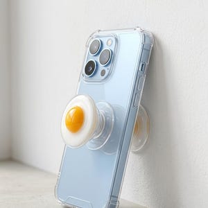 Op de afbeelding: Een lichtblauwe smartphone in een doorzichtige hoes met een telefoonhouder in de vorm van een gebakken ei. De telefoon leunt tegen een witte muur. De telefoonhouder heeft een witte rand en een gele kern.